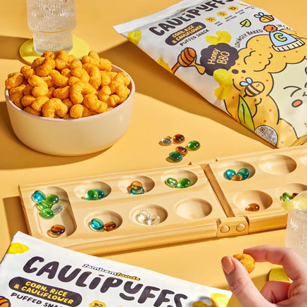 Bowl of CauliPuffs Honey BBQ on a yellow table with a bag of puffs, a glass of water, and a board game with colorful marbles