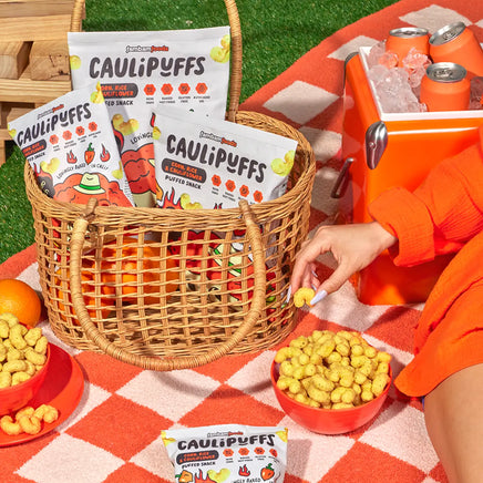 Wicker basket filled with bags of CauliPuffs Habanero Ranch on a picnic blanket, with a bowl of puffs, an orange, and a cooler with drinks